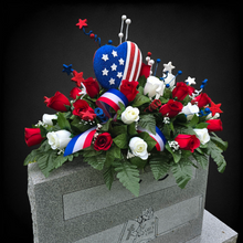 Load image into Gallery viewer, This headstone saddle is a patriotic cemetery decoration with grave flowers including red & cream roses and large American flag heart.